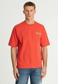 T-shirt rossa in cotone con maniche corte, girocollo, e un piccolo dettaglio di testo bianco stampato sul petto. Indossata con jeans blu strappati.