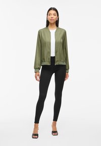 Olivovo zelená saténová bomber bunda s ribovanými manžetami a lemom, nosená cez bielu košeľu a čierne legíny, s predným zipsom a bočnými vreckami.