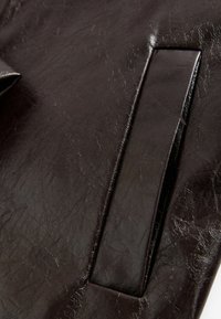 Matière en cuir synthétique marron avec une surface texturée, présentant un détail de patch rectangulaire. Fini lisse avec des motifs plissés sur toute la surface.