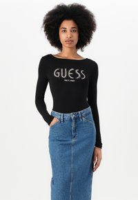 Maglietta nera a manica lunga con la scritta "GUESS EST.1981" in lettere metalliche, abbinata a una gonna in denim blu a vita alta con tasche e chiusura a bottone.