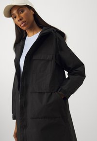 Veste longue noire avec capuche, deux poches avant et fermeture éclair. Fabriquée en tissu léger avec une finition mate.