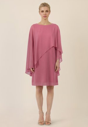 Kobieta stojąca, ubrana w różową sukienkę do kolan z asymetrycznymi przezroczystymi rękawami w formie peleryny oraz beżowymi szpilkami na kostkę na jednolitym tle.