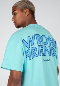 Camiseta de algodón de color turquesa claro con el texto en azul bold "AMIGOS INCORRECTOS" en la parte posterior y "CLOTHING CO." en un texto más pequeño debajo.