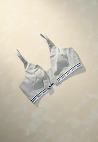 Šedý bavlnený bralet s nastaviteľnými popruhmi, ktorý má biely pás s logom "NEXT". Hladká textúra a jednoduchý dizajn.