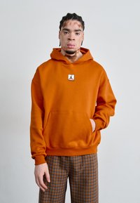 Haut à capuche orange avec une poche kangourou et un petit logo au centre ; poignets et ourlet côtelés ; associé à un pantalon à carreaux marron.