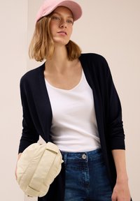 Marineblauer langer Cardigan, weißes Tanktop, blaue Jeans und eine cremefarbene Umhängetasche. Akzentuiert mit einer hellrosanen Mütze. Glatte Texturen und lässiges Design.