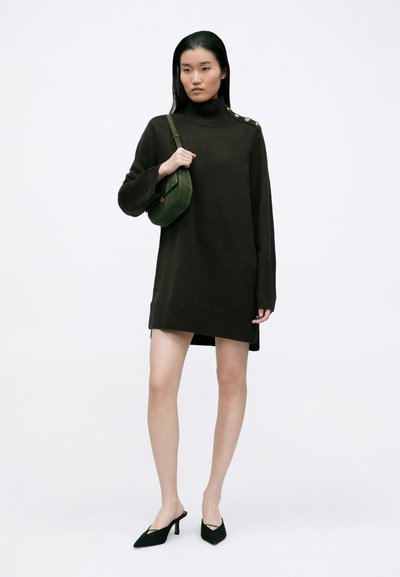 Σκούρο πράσινο πλεκτό φόρεμα με ribbed μανσέτες και στρίφωμα, διαθέτοντας ψηλό λαιμό και κουμπιά στους ώμους. Μαύρες μυτερές μπαλαρίνες και πράσινη τσάντα ώμου.