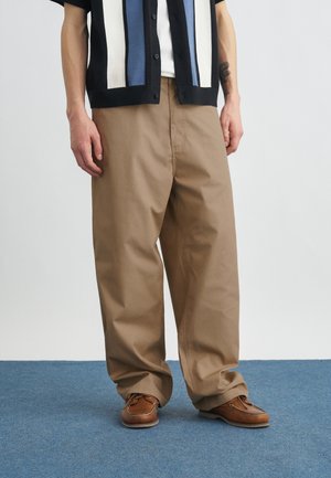 Hombre vestido con pantalones tan sueltos, zapatos náuticos marrones y una camisa de manga corta con rayas verticales en negro, blanco y azul, de pie sobre una alfombra azul.