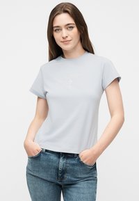 Νέα γυναίκα με μακριά καστανά μαλλιά φοράει ανοιχτό μπλε κοντομάνικο πουκάμισο με διακριτικό λογότυπο "GUESS", σε συνδυασμό με μπλε τζιν, με τα χέρια στις τσέπες.