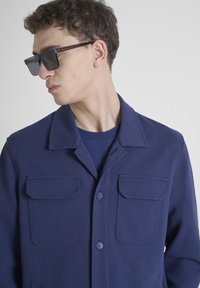 Chaqueta de botonadura en azul marino con dos bolsillos en el pecho, un cuello y una tela texturizada. El modelo lleva gafas de sol oscuras y una parte superior a juego por debajo.