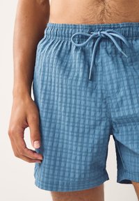 Vaaleansiniset uimashortsit, jotka on valmistettu teksturoidusta kankaasta. Shortsien vyötäröllä on kiristysnauha ja pystysuorat raidat tuovat lisäyksityiskohtia.