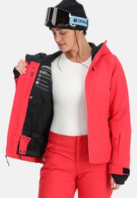 Helle rote Skijacke mit schwarzen Akzenten, mit einem Reißverschluss vorne, artikulierten Ärmeln und wasserabweisendem Material. Futter ist innen sichtbar.
