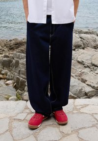 Lösa mörkblå jeans med kontrasterande vita sömmar, i kombination med röda sneakers på en stenlagd yta nära klippigt vatten.