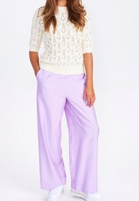 Knitad krämfärgad tröja med korta ärmar och öppen mönstring, kombinerad med löst sittande lavendelfärgade byxor och vita sneakers. Avslappnad outfitdesign.