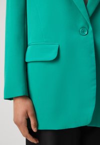 Türkise Blazerjacke mit einem einzigen Knopf, Reverskragen und Brusttasche. Glatter Stoff mit einer strukturierten Passform.