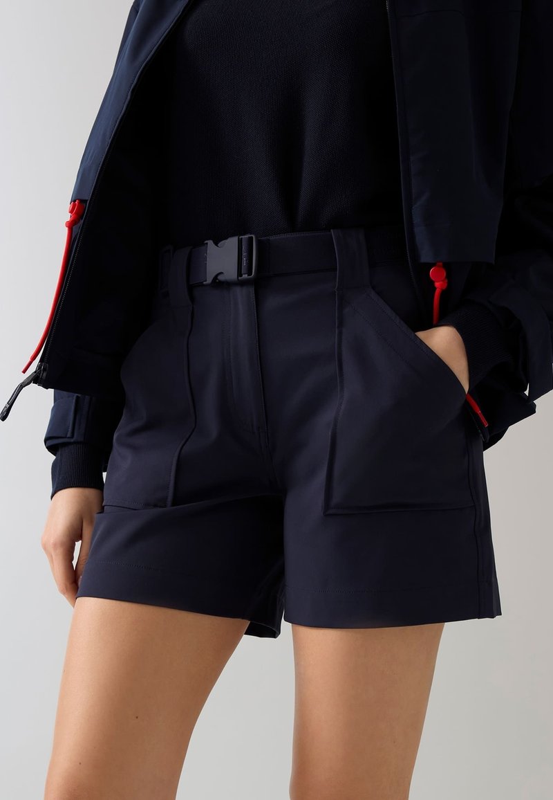 Femme portant un short noir taille haute avec des poches utilitaires et une ceinture, associé à un haut noir et une veste bleu marine avec des cordons rouges sur la fermeture éclair.