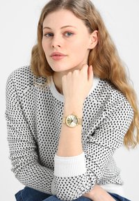 Gold watch with a metallic band, round face, and textured dial. The wearer has wavy hair and is dressed in a patterned black and white sweater.