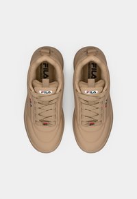 Beige sneakers med en jevn tekstiloverdel, rund tå, snøring, og Fila-merke på tungen og siden med røde og blå detaljer.