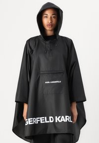 Svart vattentålig poncho med huva, featuring en framficka och fet vit "KARL LAGERFELD" text vid nederkanten. Slät, lätt material.