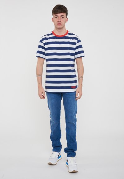 T-shirt a righe blu navy e bianche con colletto rosso, abbinato a jeans azzurri chiari e sneaker bianche. Branding di rilievo sulla maglietta.