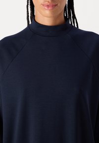 Tummansininen paita, jossa on korkea kaulus, valmistettu sileästä kankaasta. Sisältää raglan-hihat ja rento leikkaus. Yksinkertainen, minimalistinen muotoilu.