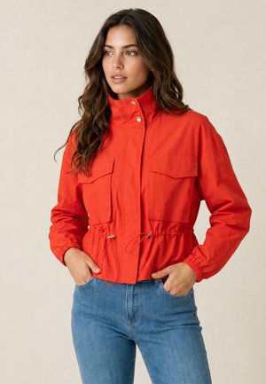 Donna con i capelli castani lunghi che indossa una giacca rossa brillante con tasche frontali e jeans blu, in piedi con le mani nelle tasche, guarda di lato.