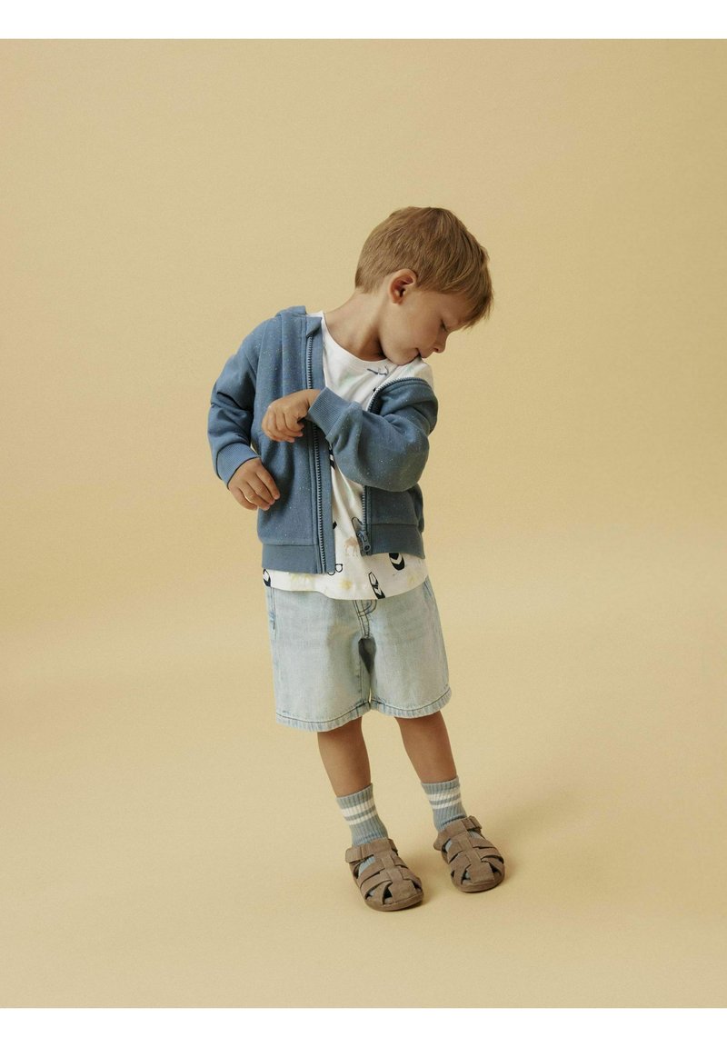 Sudadera azul con cremallera sobre una camiseta blanca con un patrón, pantalones cortos de mezclilla azul claro y sandalias beige, con un fondo neutro.