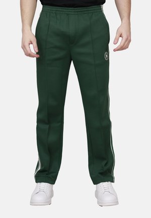 Uomo che indossa pantaloni da tuta verde scuro con strisce bianche sui lati e sneakers bianche, in piedi contro uno sfondo semplice.