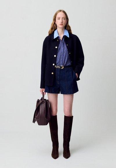 Abrigo navy con botones dorados, camisa de cuello azul, shorts de denim y botas de ante marrón hasta la rodilla. Bolso marrón con rayas verticales.