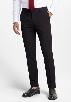 Mann trägt schwarze, maßgeschneiderte Anzughose, schwarze Leder-Loafer, weißes Hemd und rote Krawatte, steht mit einer Hand in der Tasche.