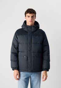 Νεαρός άνδρας φορώντας ένα σκούρο μπλε επενδυμένο μπουφάν με κουκούλα και καουτσούκ σταυρωτό, σε συνδυασμό με ανοιχτόχρωμα μπλε τζιν, στηρίζεται σε έναν απλό φόντο.