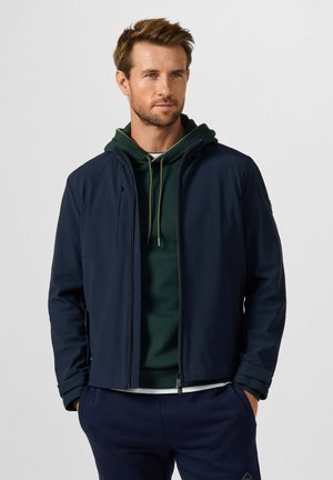 Man met een donkerblauwe jas over een groene hoodie met trekkoorden, staand met handen in de zakken tegen een effen lichte achtergrond.