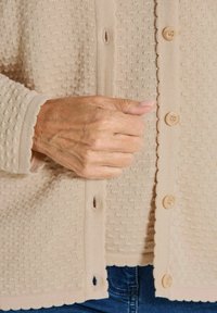 Beige strukturierter Cardigan mit Knopfschluss, ausgestattet mit einem gewellten Rand, kleinen erhabenen Mustern und einem weichen, gestrickten Stoff.