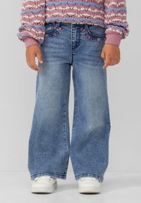 Blaue Denim-Jeans mit weitem Bein, verziert mit Blumenmotiven in der Nähe der Taschen, kombiniert mit weißen Turnschuhen. Strukturiertes, lässiges Design.