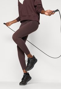 Burgundy színű leggings, texturált anyaggal és varrási részletekkel, fekete sportcipőkkel párosítva. A figura egy ugrálókötéllel áll.