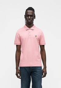 Jaunas vyras su pintomis plaukais, dėvintis rožinę trumpomis rankovėmis polo marškinėlį su geltonu siuvinėtu logotipu ir mėlynais džinsais, stovintis priešais baltą foną.