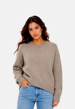 Ung kvinde med bølget brunt hår iført en løs beige sweater og lyseblå jeans, poserer med den ene hånd i lommen.