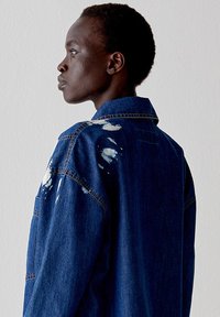 Giacca di denim blu scuro con dettagli rovinati e colletto a punta. Presenta una vestibilità morbida e cuciture visibili.