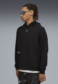 Mand med dreadlocks bærer en sort Puma hoodie, mønstrede sorte bukser og grå solbriller med gule arme, stående mod en grå gradientbaggrund.