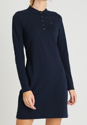 Vrouw draagt een marineblauwe polo jurk met lange mouwen, knoopsluiting en een klein logo op de borst, staand tegen een effen achtergrond.