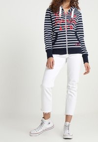 Sudadera con cremallera a rayas en azul marino y blanco, con bolsillos frontales, texto rojo, capucha ajustable, combinada con jeans blancos remangados y zapatillas blancas.