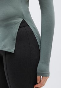 Hosszú ujjú felső tompa zöld pamutkeverékből, oldalán hasítékkal, fekete, testhez simuló leggingsszel párosítva. Sima textúra varráselemekkel.