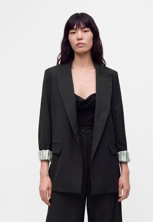 Vrouw met lang donker haar die een zwarte blazer draagt met opgerolde gestreepte manchetten en zwarte hoge taillebroeken tegen een effen achtergrond.