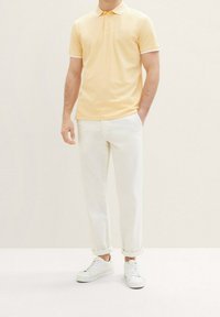 Polo shirt en jaune doux, avec un col rayé et deux boutons. Associé à un pantalon clair retroussé et des baskets blanches.
