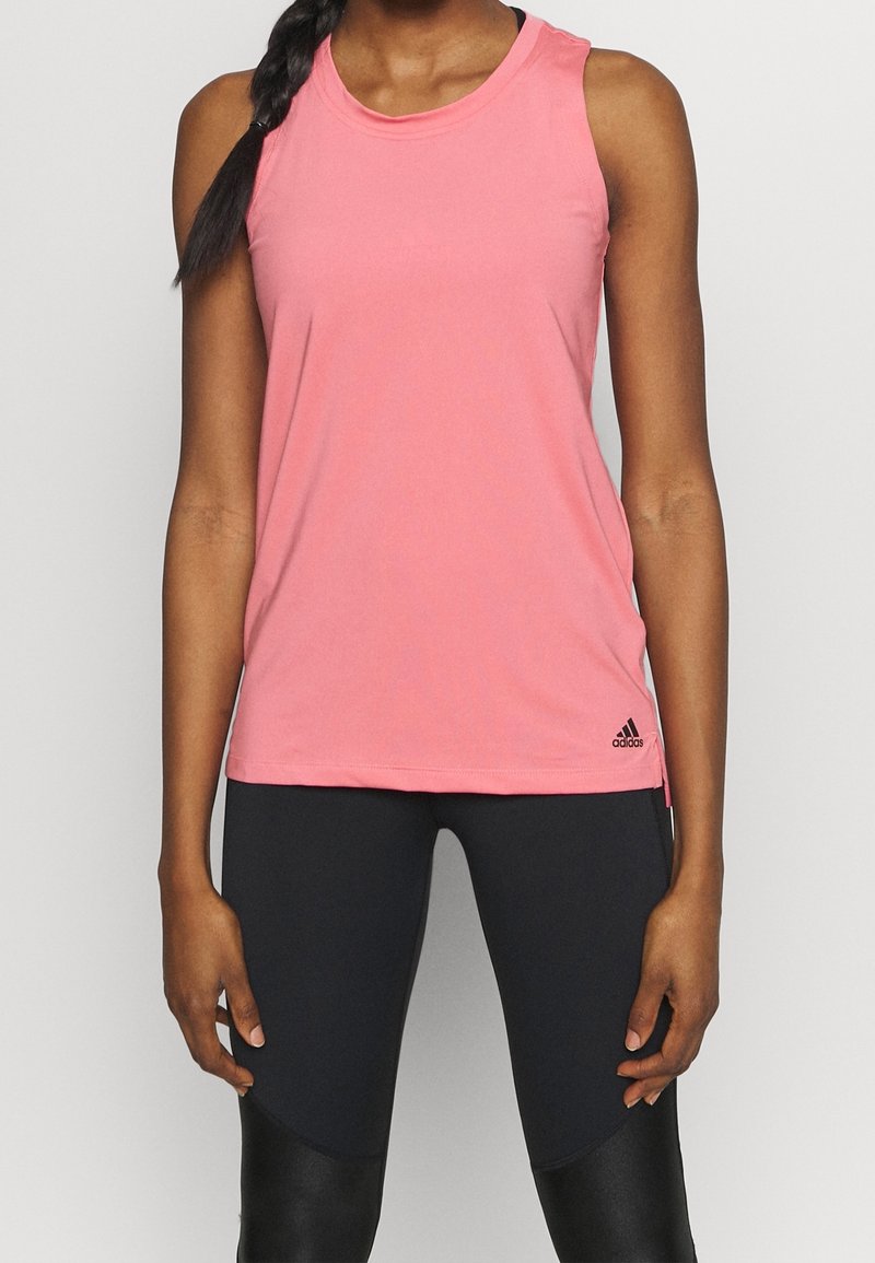 Haut de sport rose sans manches avec col rond et fentes latérales, fabriqué en tissu respirant. Présente un logo Adidas noir à l'ourlet.