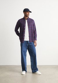 Camisa de cuadros en rojo oscuro y azul, camiseta blanca debajo, jeans azules holgados y zapatillas blancas, complementado con una gorra azul marino.