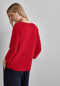 Top rosso a coste con maniche lunghe, scollatura ampia e spalle cadute, abbinato a pantaloni blu scuri. Tessuto liscio e vestibilità comoda.