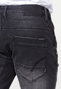 Črne jeans hlače imajo eleganten dizajn z teksturirano površino. Vključujejo zadnje žepke in blagovno označbo iz usnja na pasu.