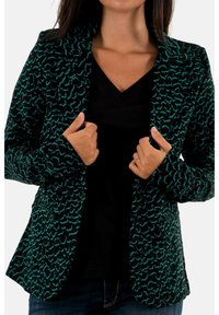 Blazer à motifs noirs et bleu sarcelle avec une coupe ajustée, des revers, et une fermeture par boutons. Porté sur un haut noir à col en V avec une finition texturée.