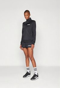 Czarny sportowy dres z kurtką zapinaną na zamek i krótkimi spodenkami. Zdobiony haftowanym logo, zestawiony z czarno-białymi paskowanymi skarpetkami i trampkami.